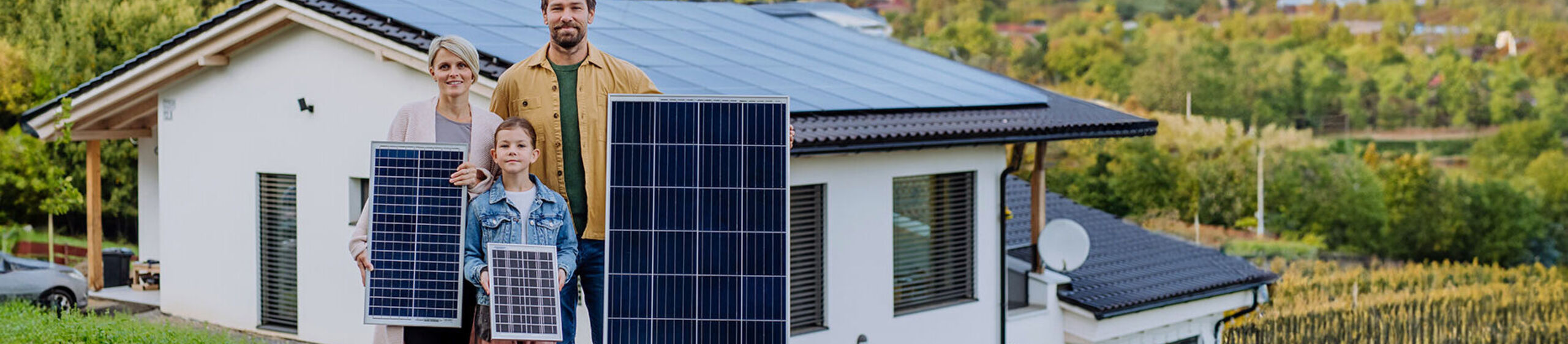 Ehepaar mit Kind steht vor einem Haus, jeder hält ein Solarpanel