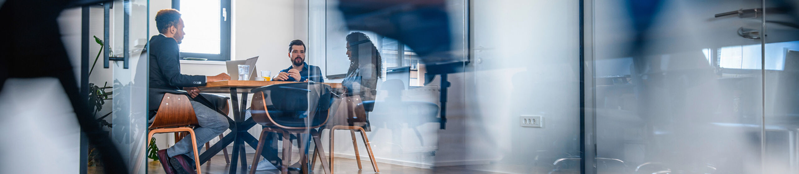 Drei Personen sitzen in einem mit Glaswänden abgetrennten Büro an einem Tisch