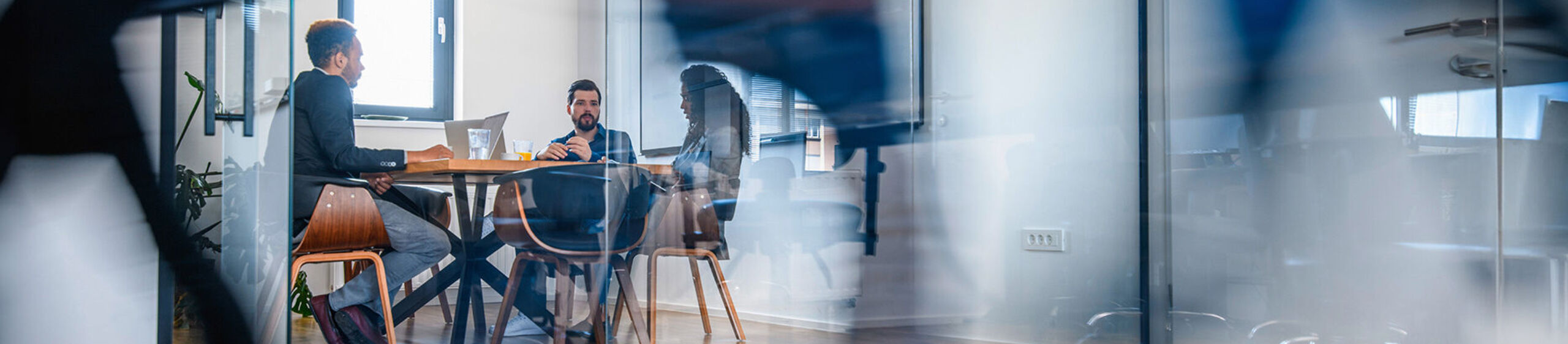 Drei Personen sitzen in einem mit Glaswänden abgetrennten Büro an einem Tisch